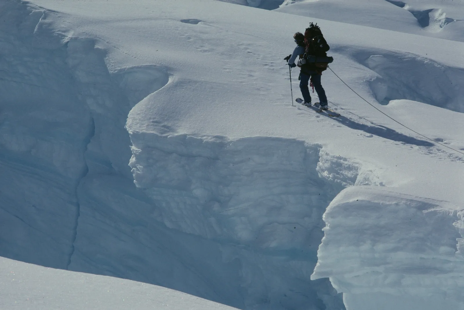 Roped mountaineer on skis skirting a deep crevasse on a glacier, the rope extending out of frame toward an unseen partner