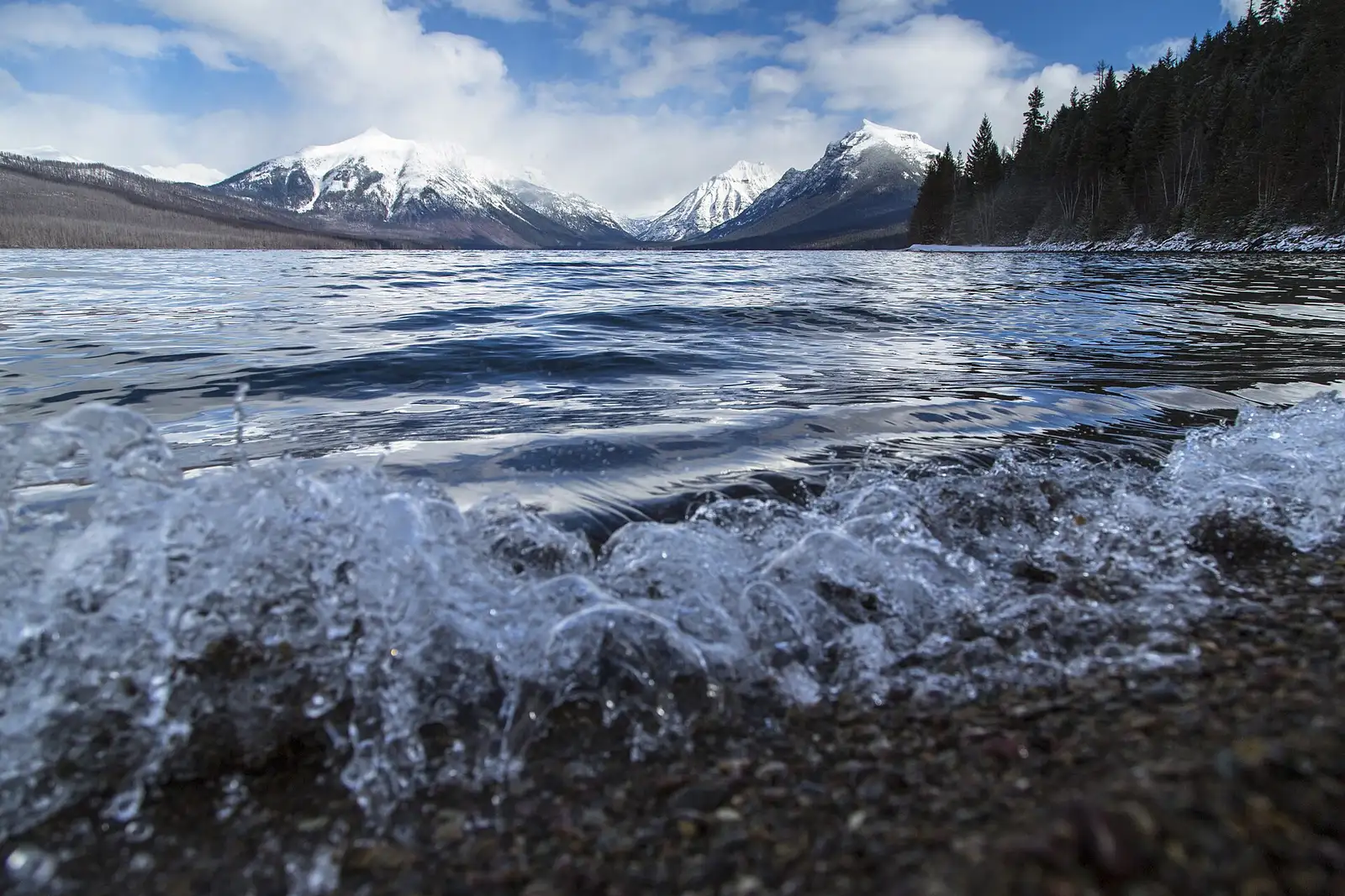 A wave breaking into crystalline spray on a lakeshore, facing snow-capped peaks under a cloudy sky